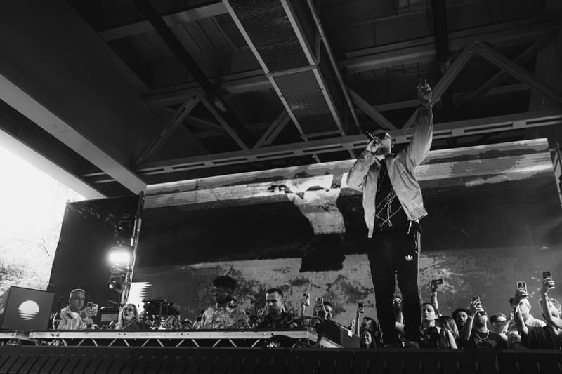 Ali Love with his arm raised on a festival stage under a steel roof, in black and white