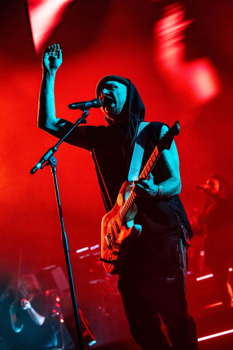 Ali Love singing with a guitar, hood up and one hand raised, under red light
