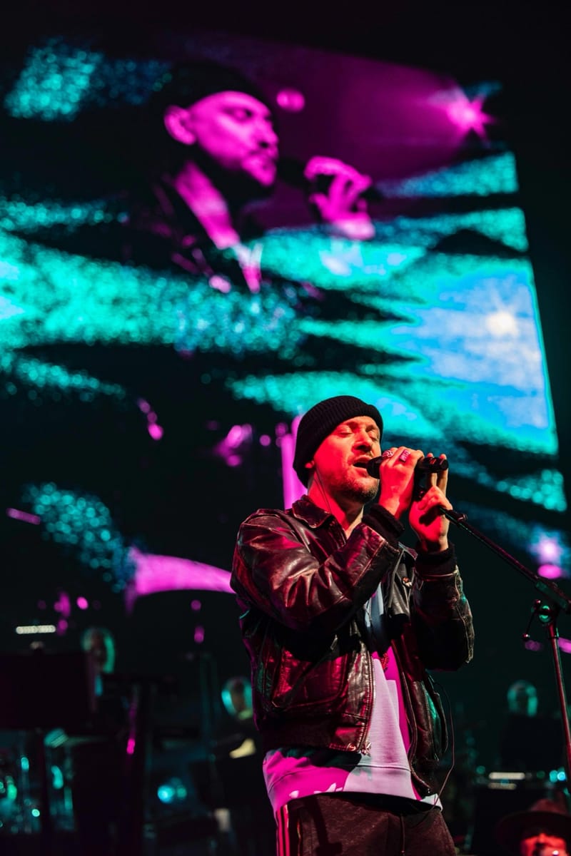 Ali Love in a beanie and leather jacket, singing in front of a screen of pink and teal light