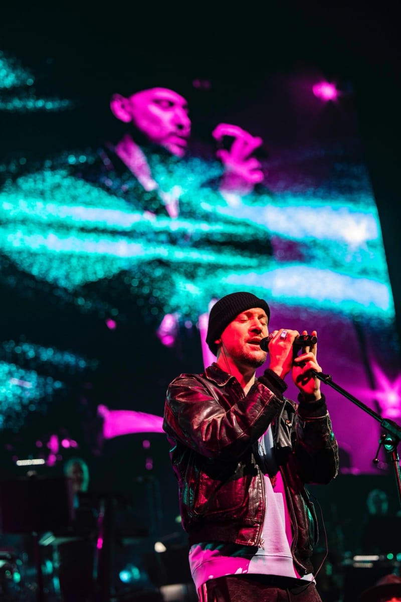Ali Love in a leather jacket, both hands on the microphone, in front of a pink and teal screen