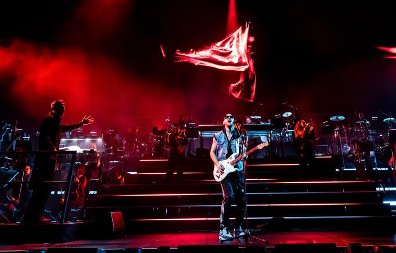 Ali Love with a guitar at the front of an orchestral stage, under red lights