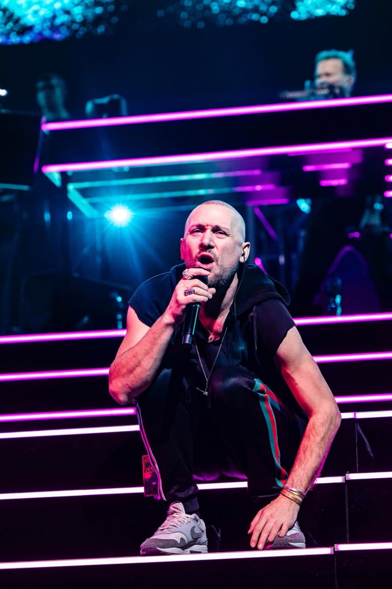 Ali Love crouching at the front of the stage with a microphone, under pink and white lights